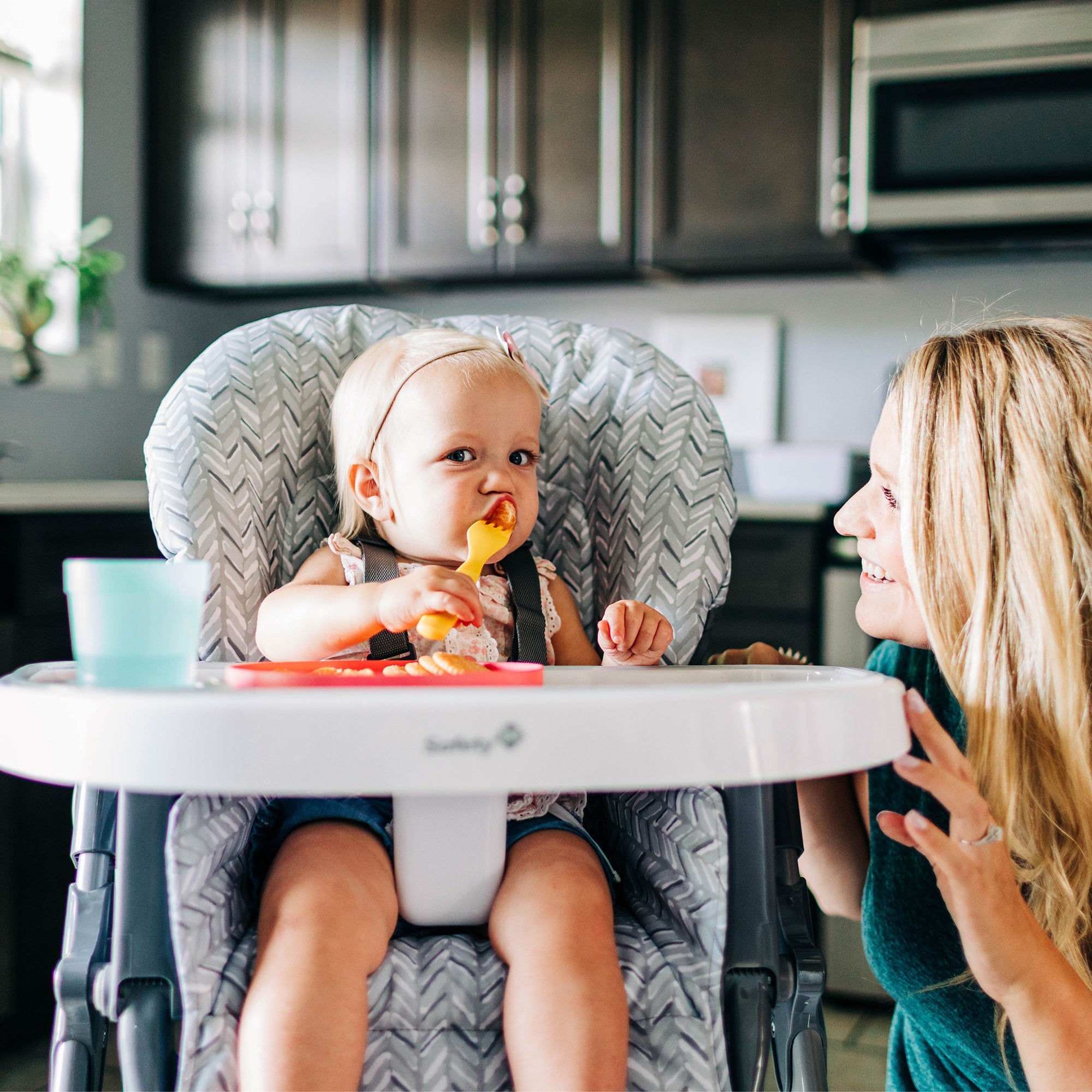 toddler girl in high chair holding fork with mom nearby