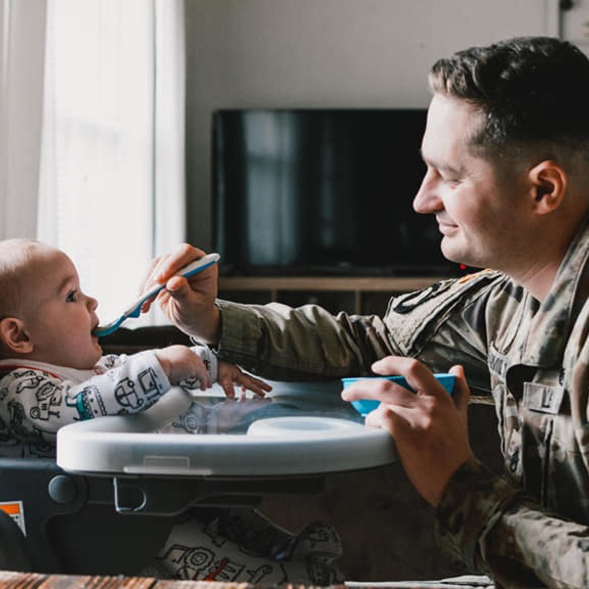Man in army uniform smiling while feeding baby in high chair